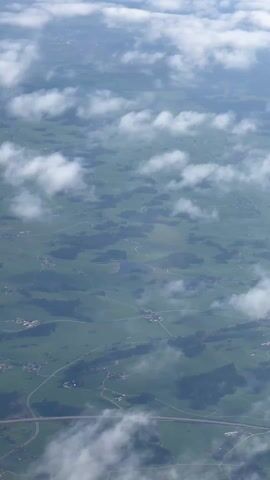 The video shows a landscape of green fields, trees, and scattered buildings from an aerial perspective, partially obscured by clouds. The landscape is crisscrossed with roads.