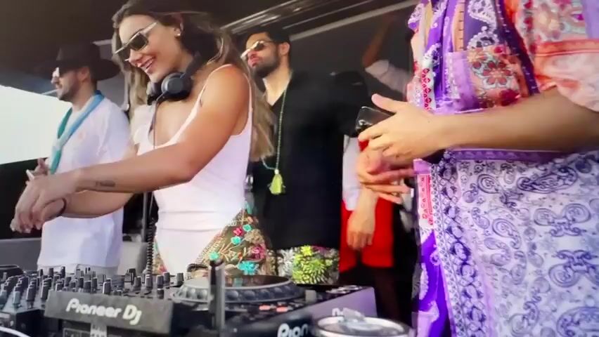 A woman DJ is mixing music on a Pioneer DJ setup at a party. Several people are around her, some dancing and others watching. The background is out of focus. The scene then shifts to an aerial view of a body of water with two boats. One boat is closer to the shore and appears to be moving, leaving a wake. The other boat is further out at sea. The water is blue and the coastline is visible in the background.