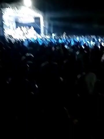 At night, a large crowd of people watches a stage performance and a projected image on a large screen. The stage is lit with bright lights, and a band is visible performing. The projected image on the screen appears to be a video or movie. The crowd is dark, with only the stage and screen illuminating them. Some people in the crowd are holding up their hands. The overall atmosphere suggests an outdoor concert or festival.