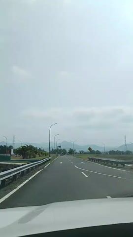A car is driving down a two-lane road with a white median barrier on the left. The road is lined with streetlights and palm trees on the left. Mountains can be seen in the distance. The sky is cloudy.