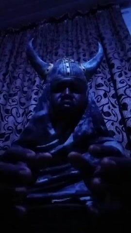 A person wearing a horned helmet and draped in a mottled blue covering sits in front of patterned curtains. The scene is dimly lit, giving the image a blue hue. The person's hands are visible and positioned in front of them. The background features a repetitive pattern of dark squiggles on a lighter background.