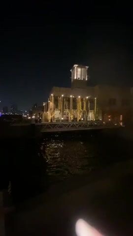 The video shows a nighttime scene of a building decorated with string lights. The building appears to be made of stone or stucco and has a tower-like structure on top. It is situated next to a body of water, possibly a canal or river, with ripples on the surface reflecting the lights. In the background, other buildings and city lights are visible, suggesting an urban setting. The overall atmosphere is festive and tranquil.