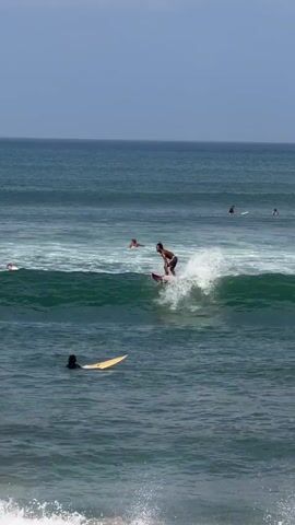 A surfer is riding a red surfboard on a green wave. The surfer is wearing dark shorts and no shirt. The surfer is positioned on the crest of the wave, carving into it. White spray is erupting from the wave around the surfboard. In the background, there are several other surfers on surfboards, some of whom are paddling in the ocean. The water is a deep blue-green, and the sky is clear and pale blue. There is a white foamy wave in the foreground, suggesting the perspective is from near the shore or from a stationary object in the water. Another surfer on a yellow surfboard is in the water to the left of the main surfer, appearing to be waiting for a wave.