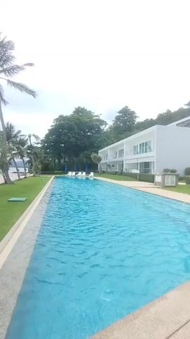 This media file captures a serene resort scene in Wichit, Thailand. The primary subjects are a long, inviting swimming pool, lush green grass, and several palm trees bordering a sandy beach and the ocean. A modern, white building with multiple balconies is visible in the background, suggesting a resort or hotel.

The activity taking place is implied relaxation and leisure, with the swimming pool being the central focus of potential enjoyment. The overall scene evokes a tranquil atmosphere, perfect for a vacation.

Notable details include the clear blue water of the swimming pool, contrasted with the vibrant green of the manicured lawn. The palm trees, with their fronds swaying gently, add a tropical ambiance. The sky appears overcast, suggesting it might be morning or late afternoon, or simply a cloudy day, which could contribute to a soft, diffused light. The location cues like palm trees, beach, and ocean strongly indicate a tropical coastal setting. There are no people visibly present, enhancing the sense of peaceful solitude.

No visible text is present in the media file.