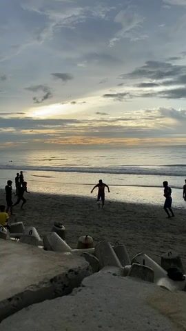 A video shows a group of people playing soccer on a beach during sunset. The sky is cloudy with rays of sunlight breaking through. The ocean waves are gentle, and the beach is covered in sand. In the foreground, there are concrete barriers. The people are silhouetted against the sunset, making it difficult to discern their features. They are actively engaged in a game of soccer, with some running and kicking the ball, while others are positioned to receive passes. The overall atmosphere appears to be one of casual recreation and enjoyment.