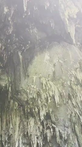 The video displays the interior of a large cave with numerous stalactites hanging from the ceiling. In the lower section of the frame, a wooden walkway is visible, with a few people standing on it. The environment appears damp and cavernous, with natural rock formations dominating the scene. The lighting is dim, suggesting an underground setting.