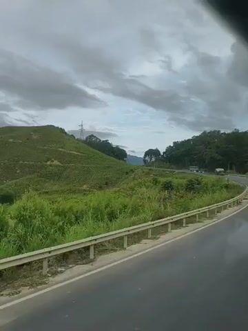 The video shows a view from a moving vehicle traveling along a paved road with a metal guardrail on the right side. The road curves to the left, offering a view of rolling green hills covered in vegetation. The sky is cloudy and appears to be overcast. In the distance, a tall transmission tower is visible on one of the hills. The overall environment suggests a rural or mountainous landscape.