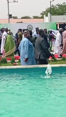 The video captures an outdoor gathering or procession in Unguwan Sanusi, Nigeria. In the foreground, a rectangular, turquoise-colored fountain actively sprays water, creating ripples on its surface.

A large group of people is visible, moving from the left side of the frame towards the right. Many individuals are dressed in traditional Nigerian attire, including long robes (agbada or caftans) and various caps. There are also men in formal suits and several individuals in what appear to be military or security uniforms, identifiable by their structured attire and caps.

A woman in a green patterned dress is a prominent figure, seen from approximately 0:01, holding up a mobile phone to record or photograph the event as she walks past the fountain. In the background, a red carpet is laid out, guiding the movement of some of the attendees, particularly those in uniform and formal wear. Behind the red carpet, a large banner is displayed, featuring a seal or logo that suggests an official capacity, possibly government-related. While text is visible on the banner, it is not clearly legible.

The scene unfolds under bright daylight, indicating sunny and clear weather. The architecture in the background appears modern, and the area includes landscaped elements like grass and shrubs, suggesting a public or institutional outdoor setting. The overall mood is orderly, with attendees observing and participating in the procession.