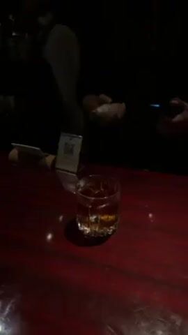 The video depicts a bartender preparing a cocktail at a dimly lit bar. The bar top is a deep red color. A lowball glass filled with a dark liquid and ice cubes sits on the bar. A small promotional stand with a QR code is also visible. The bartender, wearing a black shirt, uses a handheld torch to ignite a small piece of white material. The flame briefly flares intensely over the glass. The bartender extinguishes the torch and sets it down before picking up the glass and holding it out toward the camera.