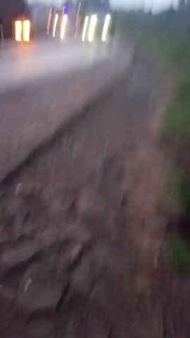 The video shows a dark, two-lane road with a vehicle approaching. The road surface appears uneven and worn. The right side of the road has a broken-up edge, with dirt and rocks visible. The video is shaky. The environment is dimly lit, suggesting it is either dusk or dawn. There are trees in the background.