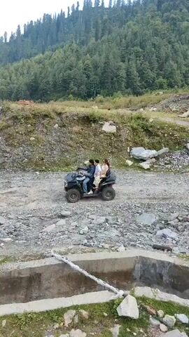 A group of people ride an ATV over a rocky, unpaved surface. The ATV is black and has three passengers. The background features a wooded hill and mountains under a cloudy sky.