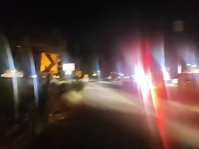 The video shows a dark road at night with a single vehicle in motion. The vehicle is moving away from the camera, and its taillights are visible. There are streetlights and the headlights of oncoming vehicles, creating lens flare. Road signs, including a yellow directional arrow sign, are visible on the side of the road. Trees and bushes line the roadside. The overall impression is of a nighttime drive on a rural or suburban road.