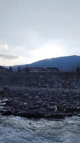 Here's a detailed description of the video:

The video shows a wide river with rocky banks. The water is turbulent and fast-flowing, appearing a light, milky blue. The river bank consists of numerous dark gray rocks of various sizes. Beyond the rocks, there's a slightly elevated area where some buildings are located, including structures with red roofs and dark supports. A white van moves on a road near the buildings. Behind the buildings, a large, dark mountain range forms the backdrop, partially obscured by clouds. The sky is overcast with bright patches, suggesting the sun is behind the clouds, and the overall lighting is somewhat muted, indicating either early morning or late afternoon.