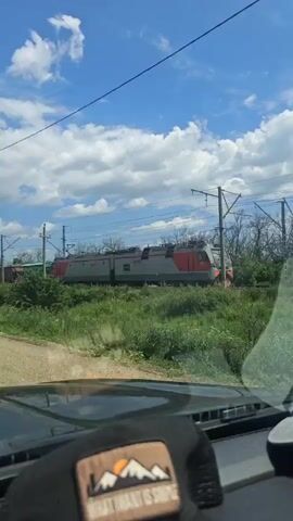 The video appears to be shot from inside a vehicle, looking out through the windshield. A train is passing by on a set of tracks to the right of the frame. The train consists of multiple freight cars, some appearing to be open-topped hoppers. Overhead power lines for the train are visible, supported by poles. A tall lamppost is also present on the left side of the frame. The sky is blue with scattered white clouds, suggesting daylight. The surrounding area is overgrown with green vegetation, including bushes and trees. The dashboard of the vehicle is visible in the foreground, with what looks like a patch or emblem on a cap resting on it.