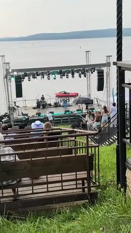 A music performance is taking place outdoors on a stage set up near a body of water. A band is visible on the stage, with a drummer and a singer. There are lights and speakers set up on the stage. In front of the stage is an audience seated on tiered benches, with some people standing on stairs to the side. The environment appears to be a grassy area overlooking the water.