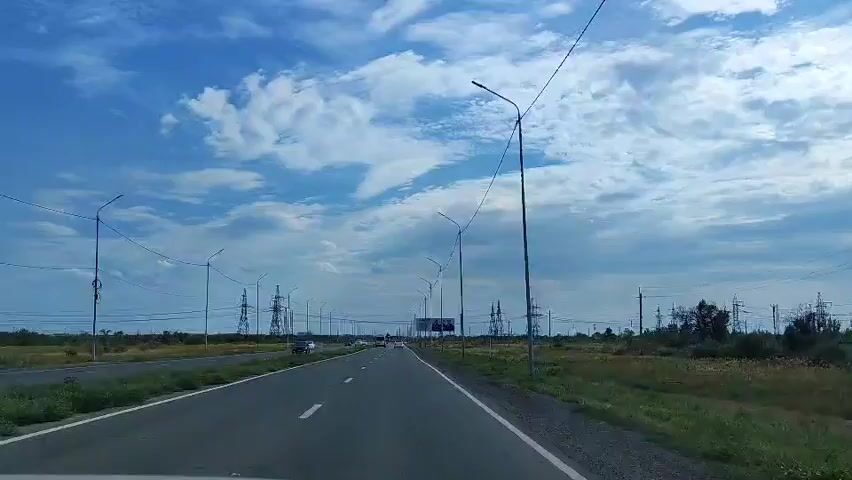 A driver's eye view of a two-lane asphalt road. The road is flanked by green grass and distant trees. Power lines and transmission towers are visible on both sides of the road. Several cars are driving in the same direction as the camera. The sky is blue with scattered white clouds.
