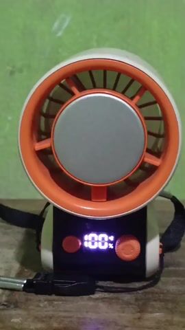 The video shows a close-up, head-on view of a portable fan. The fan is white with an orange rim around the fan opening. Inside the orange rim is a gray circular grill. The base of the fan is black and white, and it has a digital display showing "100%". There are two small red buttons on the base, one on each side of the display. A black strap is attached to the sides of the base. A black USB cable is plugged into the front of the base. The background is a blurred green wall. The fan appears to be sitting on a wooden surface. The video is very short, showing only minor movement and slight changes in lighting.