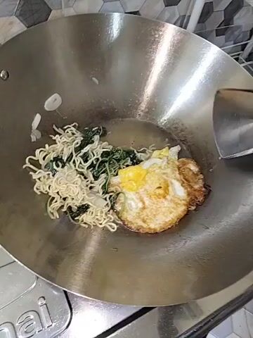 A frying pan contains instant noodles, green leafy vegetables, and a fried egg. A spatula is used to stir the ingredients in the pan, which is placed on a stovetop. The noodles are being tossed and mixed with the other ingredients. The fried egg is slightly overcooked and has a crispy edge. The vegetables appear to be wilted from the heat. The pan is made of metal and has a silver finish. The stovetop is white with black accents. The background is out of focus, but appears to be a tiled wall.