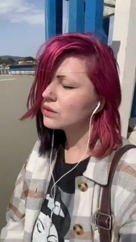 A woman with dyed pink and black hair stands outside, wearing a black t-shirt, a patterned jacket, and headphones. She has a brown purse over her shoulder. She turns her head, exposing the black dye at the root of her hair. She is on a paved surface next to a blue and white structure that looks like a part of a bridge. In the distance are parked cars. The sky is light blue.