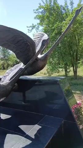 The video shows a bronze sculpture of two swans with their wings outstretched, seemingly in flight. The sculpture is placed on a dark, polished stone base. In the background, there are green trees and a clear blue sky. The camera pans across the sculpture, focusing on the details of the birds and their wings. There are some pink flowers visible in the lower right corner of the frame.