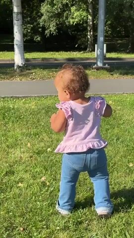 A young child, seen from behind, stands on a grassy lawn. The child has curly brown hair and is wearing a pink ruffled shirt and blue jeans. The child is facing away from the camera, looking towards the right. Trees and a paved path are visible in the background. A white van drives by on the road in the distance. The sun is shining, casting shadows on the grass.