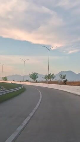 The video shows a road with lampposts and trees lining the side. The road curves to the left. There are mountains in the background, and the sky is cloudy.