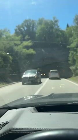 The video captures a first-person perspective from inside a vehicle driving on a road. The initial frames show the car driving on a sunny day, with trees lining the sides of the road and other vehicles visible ahead. The car then enters a tunnel, and the lighting changes dramatically. Inside the tunnel, the road surface is visible, with lane markings. Several other vehicles are seen traveling in the same direction, including a bus and cars. The tunnel walls appear to be made of rough stone or concrete. The video ends as the car continues through the tunnel, with light visible at the far end.