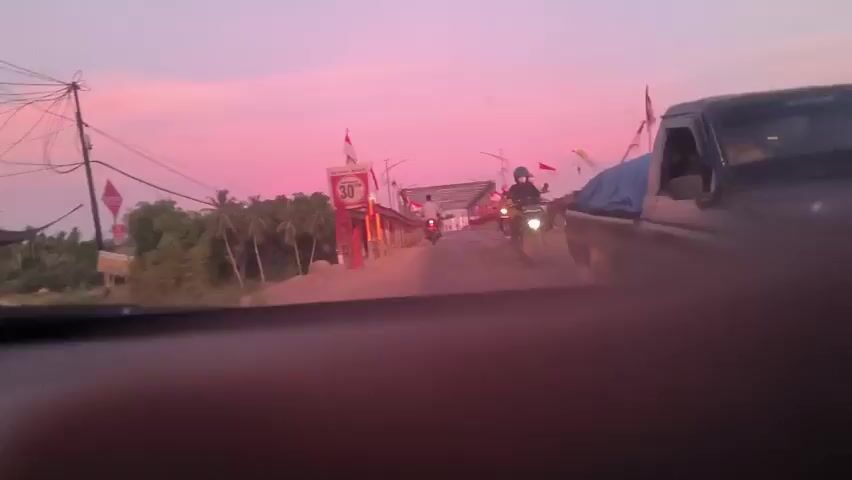 The video is shot from inside a vehicle approaching a bridge at dusk. The sky is a vivid pink. The bridge is decorated with red and white Indonesian flags and has a 30-ton weight limit sign. There are three motorcycles on the bridge heading in the opposite direction. The video is slightly blurry and the focus is initially obstructed by a finger.