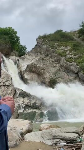 A video captures a powerful waterfall cascading over rocky terrain. The water is a murky, light brown color and churns vigorously at the base of the falls. Surrounding the waterfall are large, gray rocks and steep, eroded slopes covered in lush green vegetation. Some plastic bottles and debris are visible on the bank near the bottom of the falls. The sky appears overcast and gray. In the foreground, the hand of a person wearing a blue jacket is visible, holding the camera.