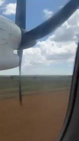 The video shows a view from an airplane window. In the foreground is part of the airplane, including the propeller spinning rapidly. Beyond the propeller, the ground is visible, appearing to be a dry, grassy plain with some trees. The sky is blue with scattered white clouds.