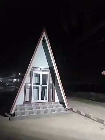 The video shows a small, triangular building at night. The building has white walls with red trim and a white door with glass panes. There are a few steps leading up to the door. The building is located in a sandy area, possibly a beach, with a streetlight visible in the background. The sky is dark and clear.