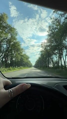 This is a first-person perspective video clip filmed from inside a car driving on a rural road. The dashboard, including the steering wheel and speedometer, is visible in the foreground. A person's hand, adorned with a silver ring, is resting on the steering wheel.

The road is paved and appears to be a two-lane highway. It is lined with lush green trees on both sides, creating a tunnel-like effect. The sun is shining brightly through the tree canopy, casting rays of light and creating a dappled effect on the road and windshield. The sky is blue with scattered white clouds.

The overall scene suggests a sunny day, likely mid-morning or early afternoon given the sun's position. The weather appears pleasant for driving. The setting is a scenic, natural environment, likely in or around Teofipilka, Ukraine, as indicated by the location context. The trees suggest a rural or semi-rural area. There are no explicit emotional cues conveyed, but the bright sunlight and scenic drive could evoke feelings of peace or enjoyment. No visible text is present in the video clip.