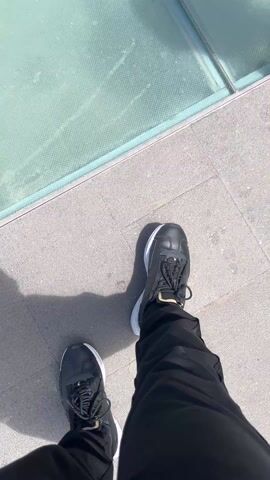 The video opens with a close-up of a person's foot in a black shoe, standing on gray pavers next to a glass panel set into the pavement. As the camera pans upward and outward, the viewer sees a glass-walled overlook along a high, rocky cliff, revealing an expansive ocean view with other islands in the distance under a blue sky dotted with fluffy white clouds. A woman in a dress and sunglasses walks into frame and looks out over the ocean.