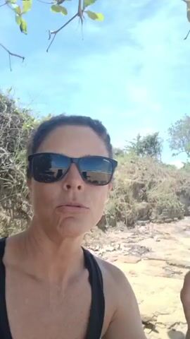 A woman with dark hair tied back, wearing sunglasses and a black tank top, speaks directly to the camera. She appears cheerful, smiling and gesturing with her right index finger. She says in Spanish, "Una caminata para que luego este desastre," which translates to "A walk so that later this disaster." The camera then pans to reveal a sunny beach scene.

The setting is a sandy beach with moderate ocean waves breaking on the shore under a clear blue sky. There is some seaweed or natural debris scattered on the sand. In the background, low coastal vegetation and a small rocky/earthen cliff are visible. The weather is bright and sunny, indicative of daytime. The overall mood is positive and energetic, despite the potentially humorous or sarcastic comment about a "disaster" following the walk. The location provided is Luquillo, United States of America, consistent with a tropical beach environment.