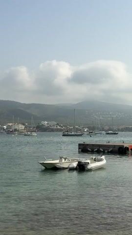 The video shows a body of water with several boats. In the foreground, two small white boats are tied together and floating. In the middle ground, there is a dock with some orange fenders. Behind the dock, several sailboats are anchored or docked. In the background, there are hills with buildings and trees. The sky is partly cloudy. A bird flies across the sky in the upper left quadrant of the frame.