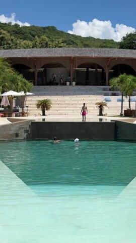 The video depicts a serene scene at a luxurious resort. A turquoise swimming pool fills the foreground, with two people swimming in it. A man in pink shorts stands on a low stone wall at the pool's edge. Behind the pool, a grand staircase leads up to an open-air building with a high wooden roof, supported by arches. Several people are seated in the shade of the building. Palm trees and other greenery surround the scene. The sky is bright blue with scattered clouds.