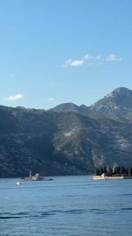 The video shows a wide shot of a bay with mountains in the background. In the foreground, there is a body of water with a small island in the middle. The island has a church on it and a few boats are sailing around it. The sky is clear and blue. The mountains are covered with green vegetation. The video is shot from a moving vehicle, as evidenced by the blurred foliage in the bottom right corner.