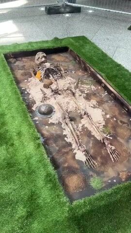 The video shows a skeleton lying in a rectangular metal container. The container is filled with wet, sandy material and is surrounded by artificial green turf. The skeleton is mostly complete, with a skull, rib cage, spine, arms, and legs visible. A metal helmet and a small tin or canister are positioned near the skeleton. The scene is indoors, with a tiled floor and metal safety barriers in the background. Signs with construction symbols are visible. There's no active movement or interaction depicted in the video.