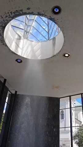 The video shows a close-up of a fountain inside a modern building. A skylight with a geometric pattern is visible in the ceiling, allowing natural light to illuminate the space. Water sprays upwards from a circular basin, creating a mist that partially obscures the lower part of the fountain. Two small blue lights are embedded in the ceiling near the skylight, and a single white light is also present. The walls are made of dark, textured stone. Large windows on the right side of the frame offer a glimpse of a white building with windows and trees outside. There are no visible people in the immediate vicinity of the fountain.