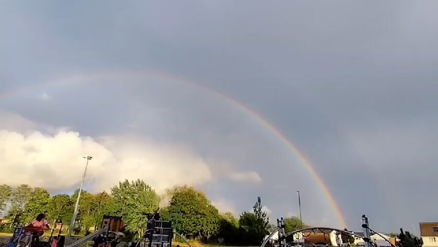 The video shows a park scene under a cloudy sky with a rainbow. In the foreground, there are children playing on playground equipment. One child is on a swing and appears to be moving. Another child is on a bicycle, also in motion. Other children are present on the ground. In the background, there are trees, streetlights, and some buildings. The rainbow stretches across the sky, with its colors faintly visible against the gray clouds. The overall atmosphere is that of a normal day at a park, with the rainbow adding a natural spectacle.