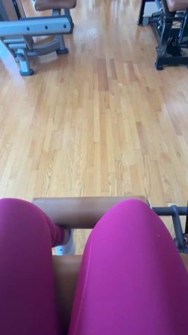 Here is a description of the video:
The video shows a first-person view of someone sitting on a leg extension machine at a gym. The person is wearing bright pink leggings and white shoes. They are positioned in front of the padded bar of the machine. The gym has wooden flooring and other exercise machines can be seen in the background. A person wearing grey pants and black shoes walks past in the background.