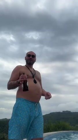 A bald, middle-aged man with a beard and sunglasses is standing outside next to a pool. He is wearing blue swim trunks with a yellow pattern and no shirt. He is holding a brown bottle in his right hand and gesturing with his left hand. The sky is cloudy and overcast. There is a body of water visible in the lower right corner of the frame, and trees and buildings can be seen in the background on a hillside. Another person is visible in the water in the lower right corner. The man appears to be speaking.