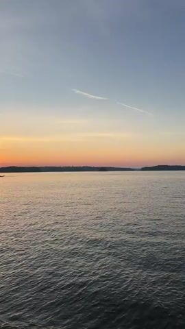 The video shows a wide body of water during a sunset. The sky is a gradient of blue and orange, with a few wispy clouds. The sun is setting on the left side of the frame, casting a warm glow over the water. The water is choppy, with small waves. In the distance, a silhouetted shoreline can be seen. The video is shot from a stationary position, likely on the shore or a dock. The overall mood is peaceful and serene.