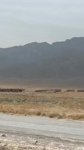 The video captures a wide shot of a desolate, mountainous landscape. A dry, grassy plain stretches towards a large, hazy mountain range under a pale sky. In the mid-ground, several small, scattered structures that appear to be made of mud or stone are visible. Some of these structures are in a state of disrepair. Further to the right, a line of what look like trucks or vehicles are parked. The foreground shows a dusty, unpaved road. The camera then zooms in on a man driving a decorated tractor. He is wearing traditional clothing and a headscarf. The tractor is adorned with colorful tassels and fabrics. The man is looking towards the camera.