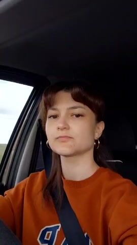A young woman with brown hair and bangs, wearing an orange sweatshirt with "GAP" written on it, is sitting in the driver's seat of a car. She is looking forward and speaking, gesturing with her left hand. Her seatbelt is fastened. The interior of the car is dark, and a glimpse of the outdoors is visible through the window.