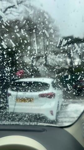 The video shows a view from inside a car, looking out through the windshield which is covered in soapy water. The focus is on the white droplets and streaks on the glass. Beyond the windshield, a blurry white car is visible, parked ahead. Other vehicles can be seen in the background, along with bare trees suggesting it may be winter. The interior of the car from which the video is shot is also visible, including a white steering wheel. There appears to be movement in the soap on the windshield.