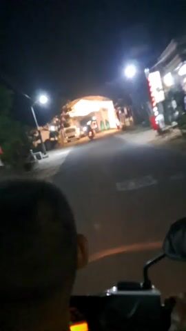 The video shows a night scene from the perspective of someone riding a motorcycle. The rider is heading towards a brightly lit white tent structure that appears to be set up over a road. There are streetlights visible along the road. Several vehicles, including a white van and a motorcycle with a rider, are present near the tent. The tent is decorated with white fabric and lights. The overall impression is that of an outdoor event or gathering taking place at night.