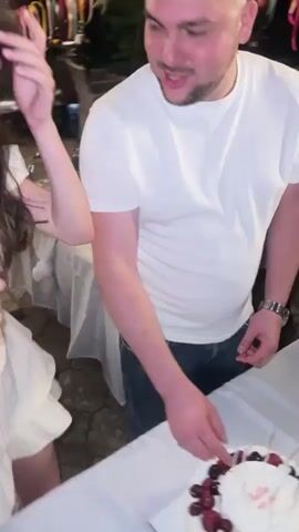 A group of people are gathered around a table outdoors at night. The table is covered with a white tablecloth and decorated with a two-tiered white cake topped with strawberries and cherries. The cake has writing on it that says "hello MY 30". A woman with long brown hair is wearing a white dress and is laughing. A man in a white t-shirt and jeans is standing next to her and appears to be cutting the cake. Other people are visible in the background, some of whom are also looking at the cake. String lights are visible in the background, indicating an outdoor setting.