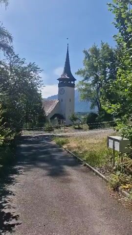 The media file shows a view of a church tower with a clock, surrounded by trees and greenery. The sky is clear and blue, suggesting a sunny day. The scene appears to be a rural or semi-rural setting, possibly a village, given the presence of the church and the natural landscape. The time of day seems to be daytime, likely mid-morning or early afternoon, judging by the bright sunlight casting shadows. There is a paved path in the foreground, leading towards the church. A mailbox is visible on the right side of the path. The architecture of the church, particularly the spire and the clock face, are prominent features. The overall atmosphere is peaceful and serene. No people are visible in the frame. No discernible text is visible in the media file, except for the timestamps which indicate the progression of time. The location context provided is Wattenwil, Switzerland, which aligns with the European architectural style of the church and the scenic, mountainous backdrop that can be inferred from the distant landscape.
