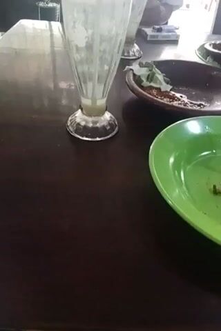 The video shows a dark brown table in what appears to be a restaurant or cafe. On the table are several used dishes and glasses. In the foreground, a green plate is visible with remnants of food. To its left, a brown plate with some food scraps and a white napkin is seen. Further back, another brown plate with food is present. There are at least three tall glasses on the table, one of which appears to be partially full of a light-colored beverage. A tissue box, likely pink and white, sits on the table. The background shows wooden chairs and a green wall with some pictures or posters. The lighting suggests an indoor setting, possibly during the day. The camera is moving, giving a pan across the table.